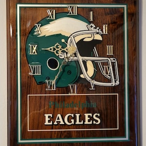 May include: A wooden wall clock featuring a green and white Philadelphia Eagles football helmet design. The clock has gold Roman numerals and hands, with the team name displayed below. The clock is framed in a dark wood.