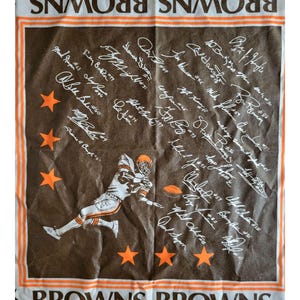 Puede incluir: Toalla del equipo Browns marr&oacute;n y blanca con un jugador de f&uacute;tbol americano, estrellas naranjas y firmas de jugadores. La palabra "BROWNS" est&aacute; impresa en naranja y blanco en los bordes superior e inferior.