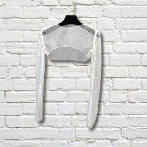 May include: A long-sleeved white bolero shrug crafted from a fine, shimmering mesh material. The garment features a cropped silhouette with a rounded neckline and long, fitted sleeves, designed to be worn over other clothing items.