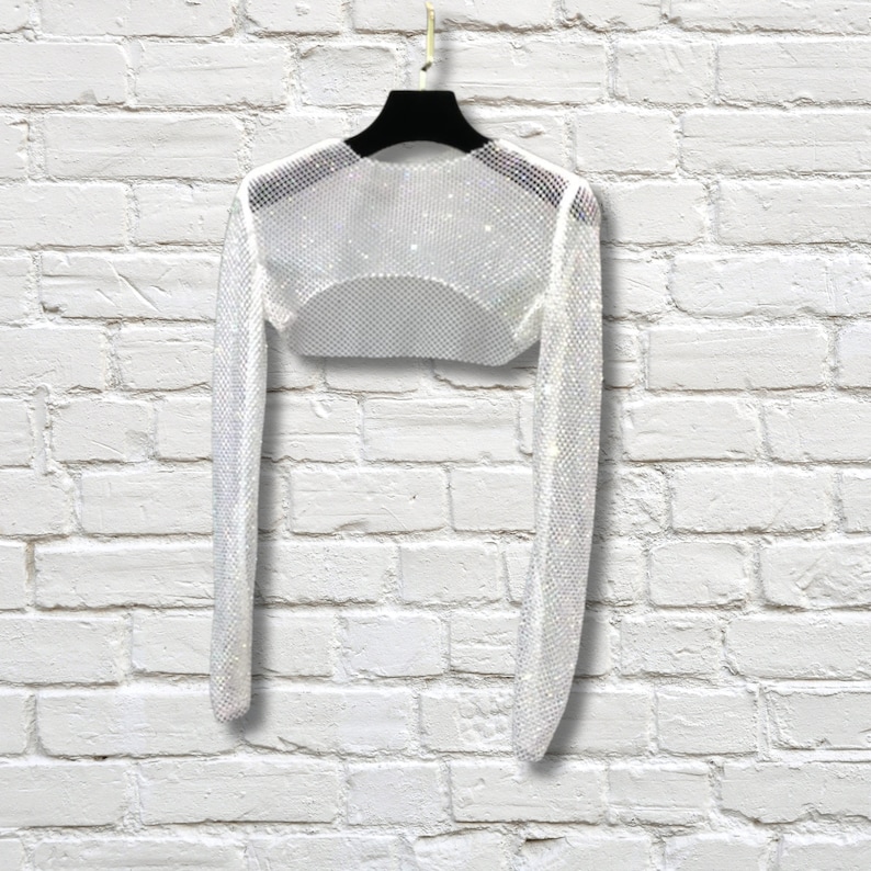 May include: A long-sleeved white bolero shrug crafted from a fine, shimmering mesh material. The garment features a cropped silhouette with a rounded neckline and long, fitted sleeves, designed to be worn over other clothing items.