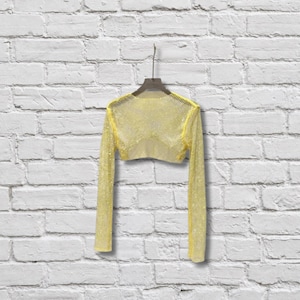 May include: A long-sleeved, cropped top made of a bright yellow, open-weave mesh fabric. The garment features a wide, rounded neckline and is displayed on a dark-colored hanger, showcasing a semi-sheer, grid-like texture throughout the entire piece.