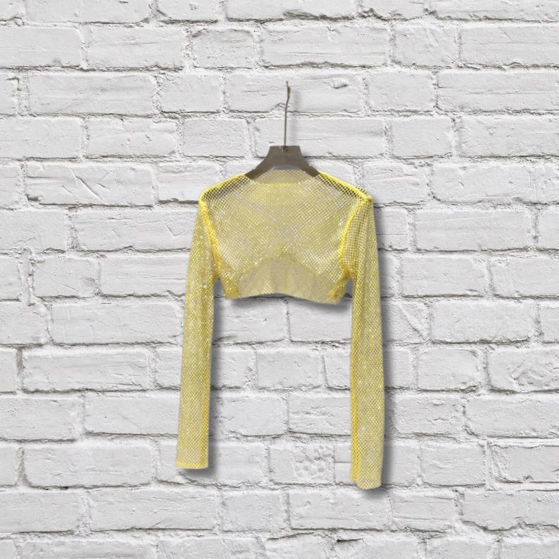 May include: A long-sleeved, cropped top made of a bright yellow, open-weave mesh fabric. The garment features a wide, rounded neckline and is displayed on a dark-colored hanger, showcasing a semi-sheer, grid-like texture throughout the entire piece.