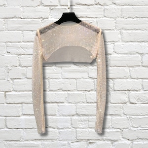 May include: A long-sleeved, cropped shrug made of a shimmering, metallic mesh fabric with a fine grid pattern. The garment features a wide, open neckline and is displayed on a black hanger, showcasing its reflective, light-catching surface.