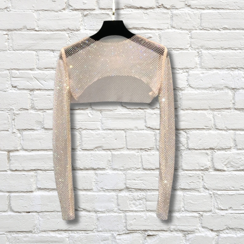 May include: A long-sleeved, cropped shrug made of a shimmering, metallic mesh fabric with a fine grid pattern. The garment features a wide, open neckline and is displayed on a black hanger, showcasing its reflective, light-catching surface.