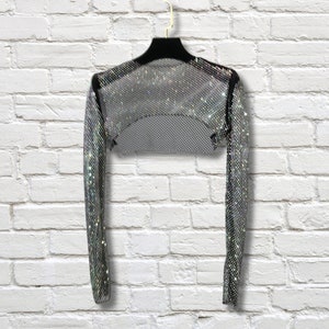 May include: A long-sleeved silver bolero shrug featuring a dense, reflective mesh construction with a shimmering, metallic finish. The garment has a cropped, open-front design with black trim along the neckline and shoulders, displayed on a hanger.