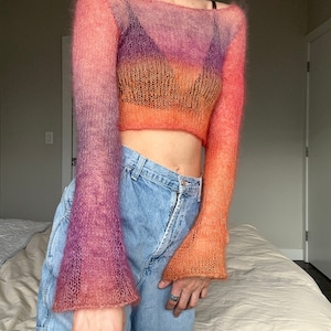 Puede incluir: Un suéter transparente de manga larga y corto con un degradado de rosa, naranja y morado. El suéter está hecho de un hilo suave y ligero y tiene un ajuste holgado.