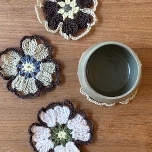 May include: Four handmade crochet flower coasters in shades of brown, cream, and blue, alongside a ceramic mug. The coasters are arranged on a wooden surface, showcasing their intricate floral design. The coasters are approximately 4 inches in diameter.