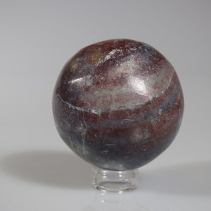 May include: A smooth, round, polished sphere of red, brown, and gray banded jasper. The sphere is displayed on a clear acrylic stand.