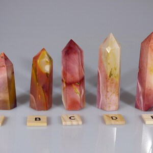 May include: Five polished, multi-colored, crystal point wands, labeled A through E, arranged in a row on a white surface.