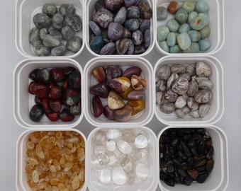 May include: Nine small white containers each filled with a different type of tumbled stones. The stones are in various colors and patterns, including grey, purple, green, red, brown, yellow, white, and black.