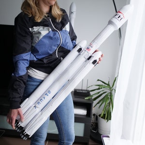 May include: A white model rocket featuring the SpaceX logo in blue lettering along the side, with Falcon Heavy branding and a small American flag decal on the upper stage. The rocket has a multi-engine base and a pointed nose cone at the top.