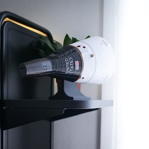 Peut inclure: Une maquette de capsule spatiale blanche et noire avec les mots "UNITED STATES" imprimés sur le côté. La capsule est posée sur une étagère noire.