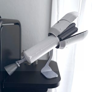 May include: A white and silver hair styling tool, resembling a rocket, is displayed on a black surface. The tool has an open section revealing black bristles. The design includes a nozzle and is supported by a silver stand.