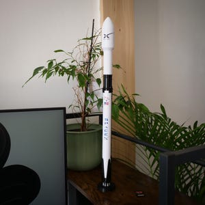 May include: A white and black model rocket featuring the SpaceX logo in blue lettering, a small American flag decal, and a black nose cone. The cylindrical structure stands vertically on a circular black base, displaying detailed markings.