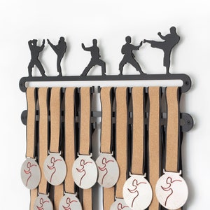 May include: Black metal medal display rack with silhouettes of karate fighters. The rack has 10 cork-backed medal hangers. The rack is mounted on a white wall.