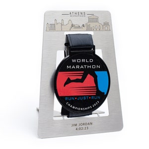 Op de afbeelding: Een zwarte en rode World Marathon-medaille met een blauwe hardloper-afbeelding, weergegeven op een zilveren standaard. De medaille leest "RUN JUST RUN CHAMPIONSHIPS 2023" en "ATHENS MARATHON" staat bovenaan.