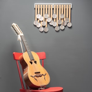 Puede incluir: Una guitarra ac&uacute;stica vintage con un dise&ntilde;o &uacute;nico se encuentra en una silla roja contra una pared gris. La guitarra tiene un cuerpo de color marr&oacute;n claro y un cuello de color marr&oacute;n oscuro con clavijas de afinaci&oacute;n plateadas. Hay 18 medallones plateados colgados en un estante de madera en la pared.