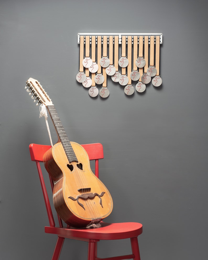 Puede incluir: Una guitarra ac&uacute;stica vintage con un dise&ntilde;o &uacute;nico se encuentra en una silla roja contra una pared gris. La guitarra tiene un cuerpo de color marr&oacute;n claro y un cuello de color marr&oacute;n oscuro con clavijas de afinaci&oacute;n plateadas. Hay 18 medallones plateados colgados en un estante de madera en la pared.