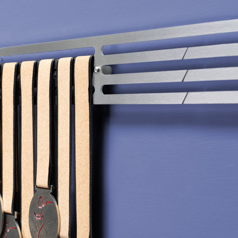 Puede incluir: Un soporte de medallas de metal plateado con cinco medallas colgadas. Las medallas son plateadas con cintas negras y beige. El soporte est&aacute; montado en una pared azul.