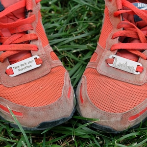 Pode incluir: Um par de tênis de corrida vermelhos com uma etiqueta prateada em cada sapato. A etiqueta do sapato esquerdo diz "New York 2019 Marathon". A etiqueta do sapato direito tem uma silhueta do horizonte da cidade de Nova York.