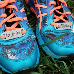 May include: A pair of blue running shoes with orange laces and a black and blue patterned design. The shoes have metal tags attached to the laces that read "Run first" and "Beer later".