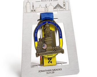 May include: A silver-toned metal display stand featuring a Boston skyline graphic, holding a marathon medal with a blue and yellow ribbon. The medal depicts a statue, a small American flag, and the text RUN TO BOSTON ROAD TO MARATHON.