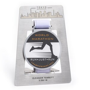 May include: A silver-colored marathon medal with a light purple ribbon, displayed on a brushed metal holder. The medal features the text "WORLD MARATHON" and "RUN JUST RUN CHAMPIONSHIPS 2023". The holder has the text "TOKYO MARATHON" and a skyline.