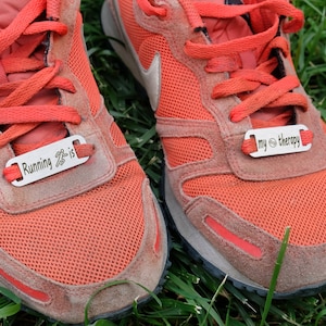 May include: A pair of red running shoes with silver shoe tags. The left shoe tag says "Running is" with a running person icon. The right shoe tag says "my therapy" with a yin yang symbol.