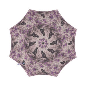 Op de afbeelding: Een paarse en zwarte paraplu met een herhalend patroon van zwarte katten en paarse bloemen.