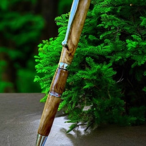 Puede incluir: Un bolígrafo de madera con un clip y detalles plateados. El bolígrafo está apoyado sobre una superficie gris con un arbusto verde en el fondo.