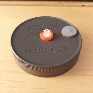 Pode incluir: Uma tampa de frasco cinza da marca Ball com as palavras "LEAK-PROOF" impressas nela. Há duas tampas pequenas, redondas, laranja e cinza na parte superior da tampa do frasco.