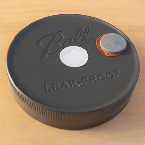 May include: Black Ball brand jar lid with a white sticker and a gray button on top. The lid says "LEAK-PROOF" in white lettering.