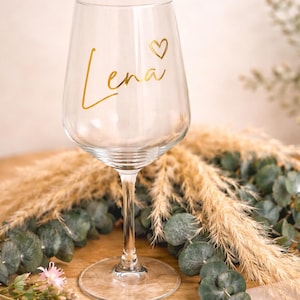 May include: A clear wine glass with a long stem and a round base. The name "Lena" is written in gold script with a heart symbol. The glass is surrounded by decorative greenery and dried grasses.