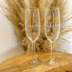 Könnte beinhalten: Zwei klare Champagnerflöten aus Glas mit goldener Beschriftung. Die linke Flöte trägt die Aufschrift "Lena" und die rechte Flöte die Aufschrift "Fabian".