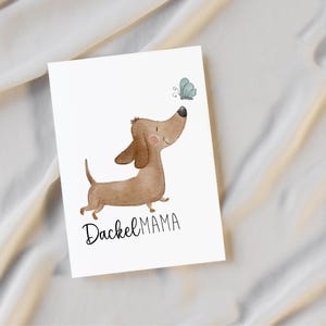 Könnte beinhalten: Eine weiße Karte mit einer Aquarellillustration eines braunen Dackel, der einen blauen Schmetterling betrachtet. Der Text "Dackel MAMA" ist in schwarzer Schrift unter dem Hund geschrieben. Die Karte befindet sich auf weißem Hintergrund.