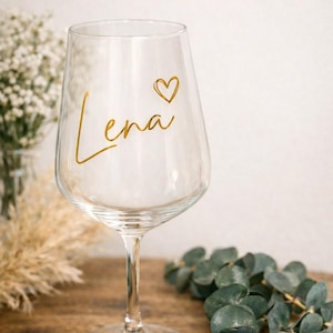 May include: A clear wine glass with the name "Lena" in gold script and a small heart. The glass is on a wooden surface, with greenery and dried flowers in the background. A personalized gift.