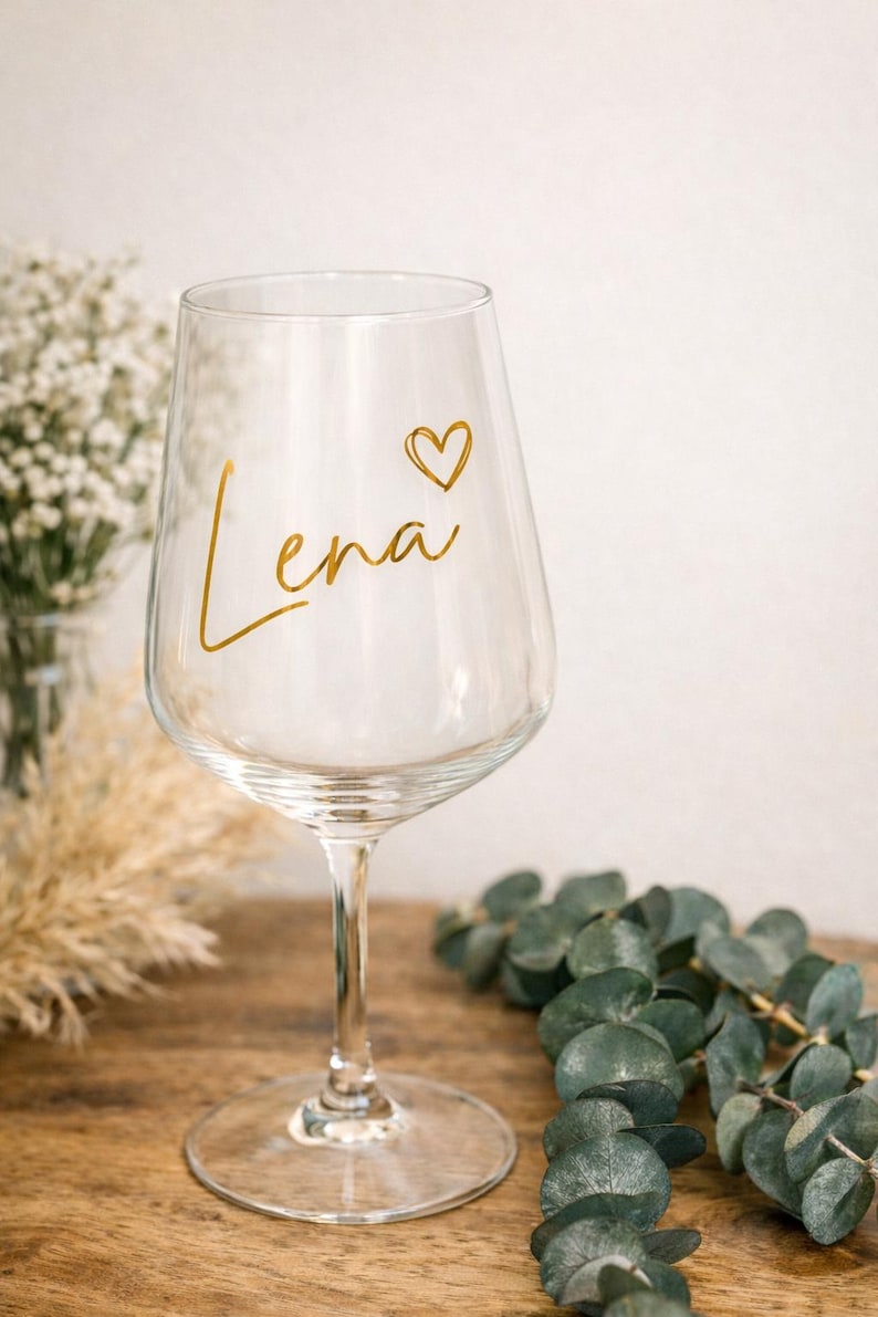 May include: A clear wine glass with the name "Lena" in gold script and a small heart. The glass is on a wooden surface, with greenery and dried flowers in the background. A personalized gift.