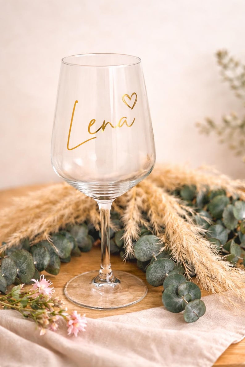 May include: A clear wine glass with a long stem and a round base. The name "Lena" is written in gold script with a heart symbol. The glass is surrounded by decorative greenery and dried grasses.