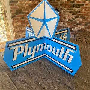 May include: A blue and white Plymouth sign with the Plymouth logo. The sign is made of wood and has a retro design. The sign is a vintage car decoration.