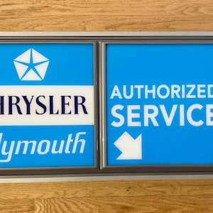 Könnte beinhalten: Rechteckiges Schild mit silbernem Rahmen, unterteilt in zwei Bereiche. Der linke Bereich hat einen blauen Hintergrund mit einem weißen Chrysler-Logo und den Wörtern "CHRYSLER" und "Plymouth". Der rechte Bereich zeigt "AUTHORIZED SERVICE" und einen weißen Pfeil auf blauem Grund.