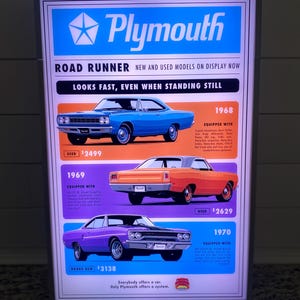 Puede incluir: Un letrero retroiluminado que anuncia los coches Plymouth Road Runner. El letrero muestra tres modelos clásicos de 1968, 1969 y 1970, cada uno en un color diferente. El letrero incluye el logotipo de Plymouth y el eslogan "Looks fast, even when standing still".