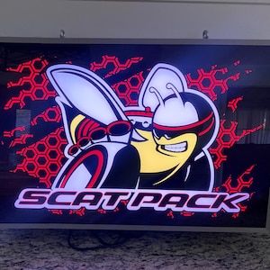 May include: A framed sign featuring the Dodge Scat Pack logo. The image shows a cartoon-style bee with sunglasses and a helmet, set against a black background with red honeycomb shapes. The word "SCAT PACK" is displayed in white.