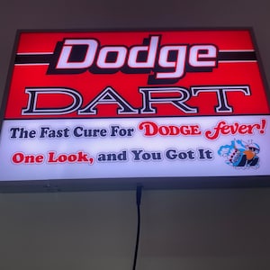 May include: Rectangular illuminated sign with the text "Dodge DART" in white and red. The sign also includes the phrases "The Fast Cure For Dodge Fever!" and "One Look, and You Got It."