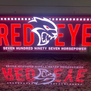 May include: A rectangular sign with the words "RED EYE" in large, red, illuminated letters. The sign also features a white logo of a snarling cat and the text "SEVEN HUNDRED NINETY SEVEN HORSEPOWER".