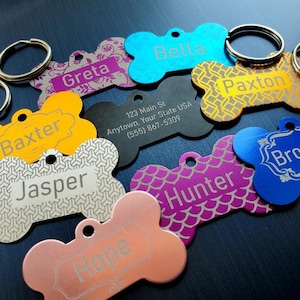 May include: A collection of colorful bone-shaped dog tags with engraved names, including Baxter, Jasper, Hope, Hunter, Brownie, Greta, Bella, and Paxton. One tag also includes an address and phone number.