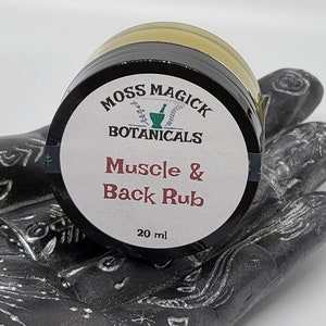 May include: A small, white jar with a black lid and a label that reads "Moss Magick Botanicals Muscle & Back Rub 20 ml".