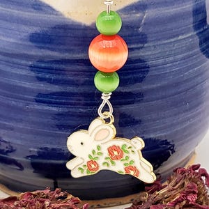 May include: A decorative charm featuring a white enamel rabbit pendant with red floral accents, suspended from a silver-toned wire loop. The charm includes two small green beads and one larger orange bead, all arranged vertically above the rabbit.