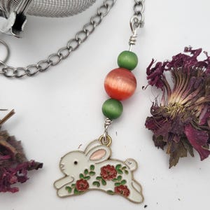 May include: A stainless steel tea infuser with a chain and decorative beads. The chain leads to a white enamel bunny charm with red floral accents. Green and orange beads are strung on the chain.