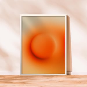 May include: Framed abstract art print featuring a vibrant orange and yellow gradient with a central circular shape. The artwork is displayed in a white frame, leaning against a wall with a wooden floor.