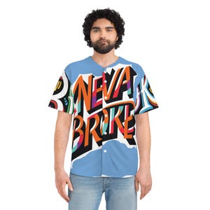 Op de afbeelding: Een lichtblauw honkbalshirt met de tekst "NEVA BROKE" in een kleurrijk graffiti-ontwerp. Het shirt heeft een knoopsluiting aan de voorkant en korte mouwen. Het ontwerp bevat oranje, paarse, zwarte en witte kleuren.